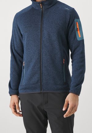 Mann trägt eine blaue Fleecejacke mit Reißverschluss und orange abgesetzten Reißverschlusstaschen sowie schwarze Hose vor einem schlichten Hintergrund.