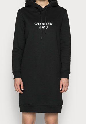 Persona con un vestido sudadera negro de manga larga con "CALVIN KLEIN JEANS" impreso en blanco en el pecho, manos relajadas a los lados.