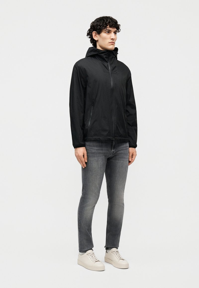 Un giovane indossa una giacca nera con cappuccio, jeans grigi sbiaditi e sneakers bianche, in piedi davanti a uno sfondo chiaro e uniforme.