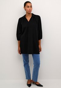 Tunique noire avec un décolleté en V, manches bouffantes 3/4 et design en tissu texturé, associée à un jean bleu clair et des chaussures noires à bout pointu.
