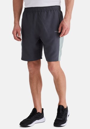 Mann trägt schwarze und graue Sportshorts sowie schwarze Sportschuhe, steht vor weißem Hintergrund.