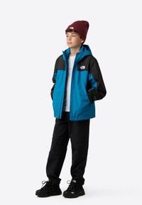 Blauw-zwarte waterdichte jas met rits aan de voorkant, capuchon en logo, gecombineerd met zwarte broek en een bordeauxrode beanie. Gestructureerde stof en sportief ontwerp.