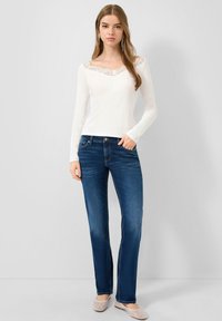 Frau steht vor einer grauen Wand und trägt ein weißes Langarmshirt mit Spitzenbesatz, dunkelblaue Jeans und beige Ballerinas.