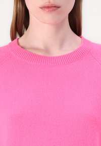 Rosa strikket genser med rund hals, synlig ribbestruktur ved kragen, og litt senket skuldersømmer.