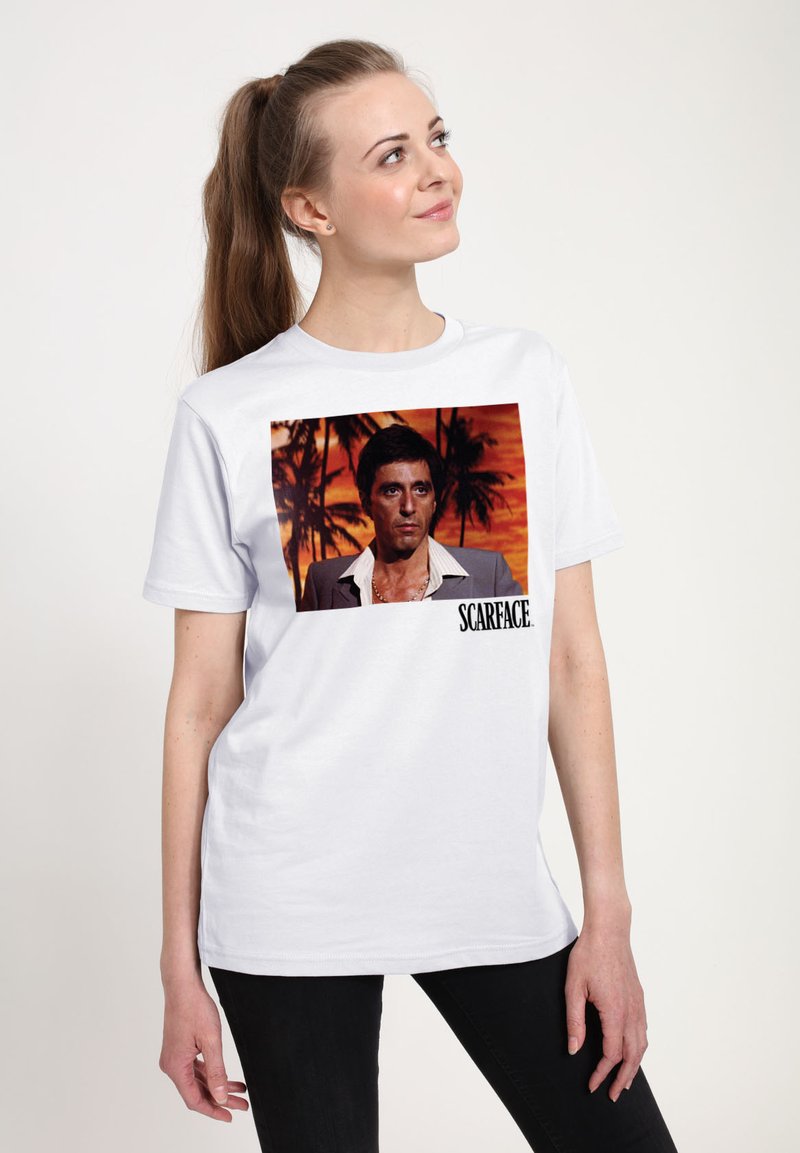 Bílý bavlněný tričko s potiskem postavy z "Scarface" na pozadí západu slunce s palmami. Černý text pod obrázkem.