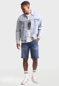 Denimjakke i lys blå, hvit t-skjorte med grønn grafik, mørkeblå denimshorts, og grå sneakers med hvite sokker med svarte striper.