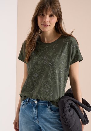 Joven mujer con camiseta de color verde oscuro con estampado y vaqueros azules, sosteniendo una chaqueta negra sobre su brazo contra un fondo beige.