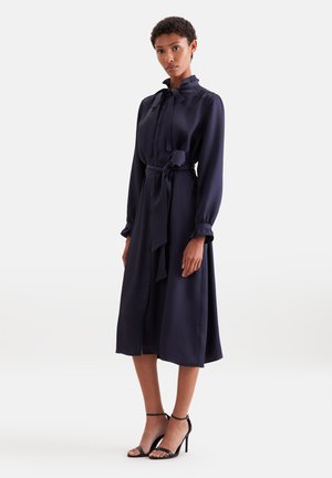 Femme portant une robe midi bleu marine à manches longues, boutonnée avec une ceinture nouée à la taille, et des sandales noires à talons hauts, debout devant un fond clair.