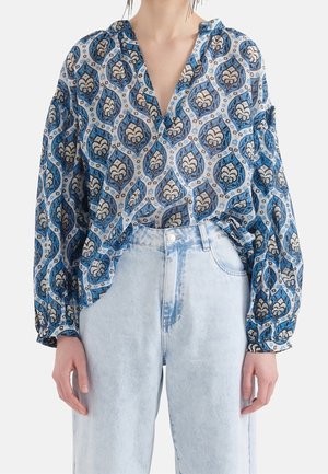 Frau trägt eine blau gemusterte langärmelige Bluse, die in hoch taillierte hellblaue Jeans gesteckt ist, vor einem weißen Hintergrund.