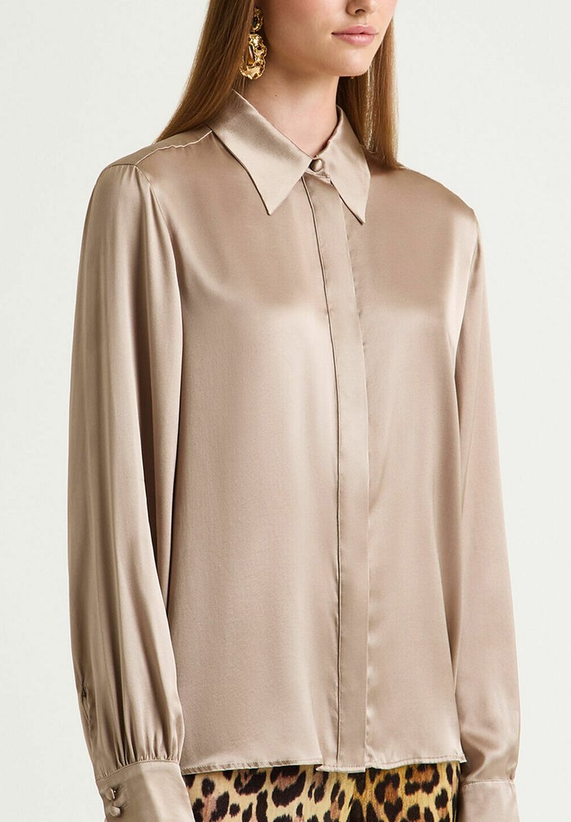 Blusa in raso beige con colletto classico, maniche lunghe a sbuffo e polsini con bottoni; caratterizzata da una texture liscia e una lieve lucentezza.