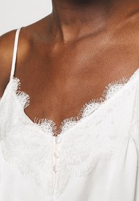 Mörkhyad person i en vit ärmlös topp med spetskant och tre tygklädda knappar i mitten.