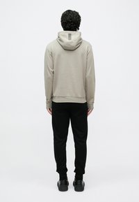 Άνδρας στέκεται γυρισμένος με την πλάτη, φορώντας μπεζ φούτερ με λεπτομέρεια ρίγες στη κουκούλα και μαύρο παντελόνι με μαύρα αθλητικά παπούτσια σε λευκό φόντο.