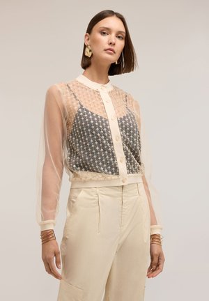 Vrouw met stijlbruin haar draagt een doorschijnende beige blouse met knopen over een zwart patroon topje en beige hoge taille broek, geaccessoriseerd.