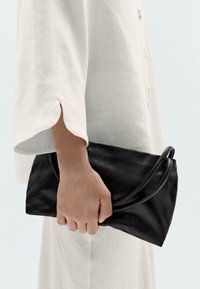 Clutch in pelle nera con cuciture curvate e accentuazioni tondeggianti, tenuto in una mano adornata con un capo a maniche lunghe di colore chiaro e leggero.