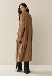 Long, loose brown cardigan made of soft fabric. Features ribbed cuffs, side pockets, and a straight hem, worn over black pants with stripes.