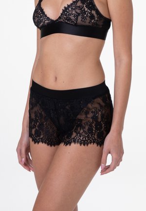 Un bralette in pizzo nero e un pantaloncino coordinato presentano un design trasparente con intricate fantasie floral e una morbida cintura in nero.