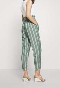 Pantalones a rayas verdes hechos de una tela suave, con una cintura elástica, bolsillos laterales y piernas ajustadas. Llevados con sandalias de cuña blancas.