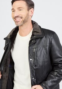 Lachende man in een wit sweater en een zwart leren jas met een gevoerde kraag, terwijl hij de jas openhoudt.