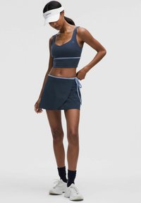 Tengerészeti sport crop top világoskék részletekkel, egy hozzá illő vállpántos szoknyával párosítva. Anyaga rugalmas; a dizájn egy testhezálló sziluettet tartalmaz.