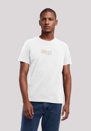 White cotton t-shirt with short sleeves featuring a subtle gold "SMILEY." text logo at the center. Model wears dark blue jeans.