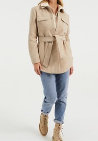 Béžová vlněná bunda s dvěma náprsními kapsami, opaskem v pase a límečkem, kombinovaná se světle modrými džíny a tanovými šn ěrovacími botami.