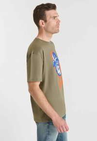 Hombre de perfil derecho vistiendo una camiseta verde olivo con un gráfico de un bulldog en azul y naranja y jeans de un azul claro, contra un fondo liso.