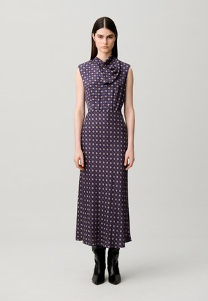 Femme aux longs cheveux foncés porte une robe midi sans manches bleu marine à motifs et des bottes noires, debout devant un fond clair uni.