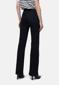 Jeans noirs évasés à taille haute, dotés de poches arrière et d'une texture lisse. Portés avec des escarpins noirs. Chemise rayée visible en haut.