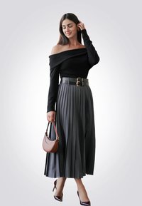 Top nero scollato con maniche lunghe, gonna plissettata grigia, ampia cintura in pelle con doppia fibbia, borsa marrone e tacchi bordeaux.