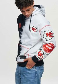 Huppari, jossa on musta-valkoinen tilkkutyömalli, Kansas City Chiefsin logot, kontrastiväriset punaiset tikkaukset ja etu-tasku.