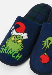 Marineblaue Plüschhausschuhe, die den Grinch in einem roten Weihnachtsmann-Hut zeigen, mit dem Text "GRINCH" auf einer Seite; die andere Seite zeigt eine rote Christbaumkugel.