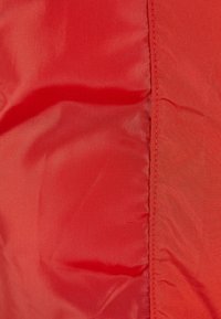Rødt stoff har en jevn tekstur med en blank finish, med en søm langs en kant. Materialet ser lett og slitesterkt ut.