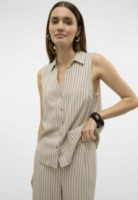 Blusa sin mangas beige con rayas verticales en azul marino, cuello y cierre de botones al frente. La tela parece suave, combinada con pantalones a rayas a juego.