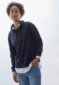 Felpa blu con cappuccio e collo a imbuto, con lacci e accessori in argento, indossata sopra una maglietta bianca, abbinata a jeans in denim azzurro chiaro.