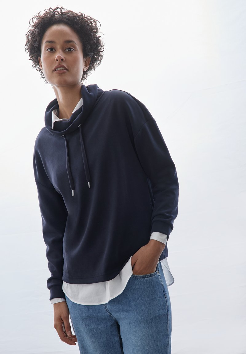 Felpa blu con cappuccio e collo a imbuto, con lacci e accessori in argento, indossata sopra una maglietta bianca, abbinata a jeans in denim azzurro chiaro.