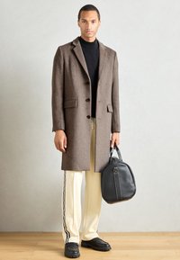 Abrigo de cuadros marrón con un corte ajustado, jersey de cuello alto negro, pantalones crema con rayas laterales y un bolso negro con acabado texturizado.