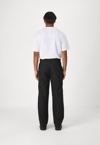 Man som står vänd bortåt, iförd en vit kortärmad skjorta, svarta byxor med bakfickor och svarta skor mot en vit bakgrund.