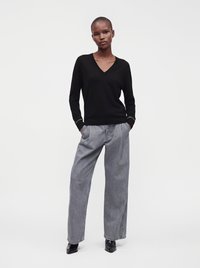 Femme debout, les mains dans les poches, portant un haut noir à manches longues encolure en V, un pantalon gris à jambes larges et des chaussures noires pointues sur fond blanc.