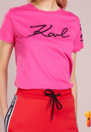 Vrouw draagt een felroze T-shirt met het Karl-logo en rode broek met trekkoord, met Karl Lagerfeld-merk aan de zijkant.