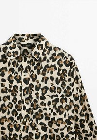 Chemise à imprimé léopard avec un col, une patte de boutonnage à l'avant et une poche avant ; présente un fond beige avec des taches noires et marron.