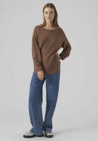 Rjav dolg rokav pulover z širokim izrezom, v kombinaciji s širokimi modrimi jeans hlačami in srebrnimi platform čevlji. Gladka tekstura, sproščen kroj.
