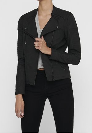 Vrouw draagt een zwarte doorgestikte motorjas over een grijs crop top, gecombineerd met een zwarte broek met hoge taille, en houdt de revers van de jas vast.