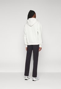 Persona con cabello trenzado, vistiendo una sudadera blanca, pantalones negros y zapatillas blancas, de pie de espaldas contra un fondo claro y liso.