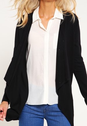 Femme portant une chemise blanche boutonnée sous un cardigan noir ample avec un jean bleu, vue du cou jusqu'aux hanches sur un fond uni.