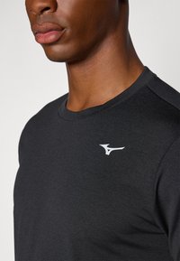 Tricou sportiv negru, cu decolteu rotund. Are un logo alb pe partea stângă a pieptului. Materialul pare neted și ușor.