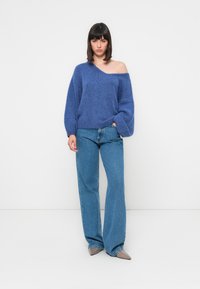 Maglione blu lavorato a maglia, grande e accogliente, con design off-shoulder, abbinato a jeans a vita alta e a campana in un tonalità di blu chiaro e scarpe grigie a punta.