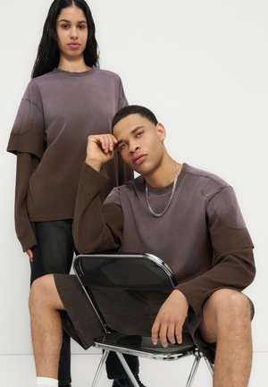 Άνδρας που κάθεται σε καρέκλα με τον αγκώνα στον αστράγαλο, φορώντας καφέ τοπ με στρώσεις και σορτς· γυναίκα που στέκεται πίσω με παρόμοιο καφέ τοπ με στρώσεις και μαύρο παντελόνι.