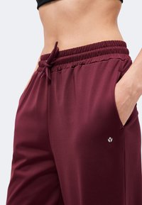 Personne portant un pantalon bordeaux à taille élastique avec cordon, une main dans la poche, recadrée au niveau du ventre sur fond uni.