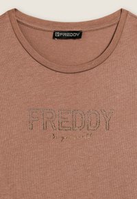 Camiseta marrón de tela suave con cuello redondo. Presenta "FREDDY" en letras con tachuelas y un texto más pequeño "sé tú mismo" debajo.