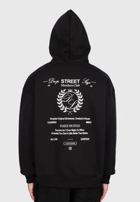 Czarna bluza z kapturem z białym napisem "Drop Street Members Club", grafiką wieńca laurowego i motywującym cytatem z tyłu, noszona przez osobę stojącą tyłem.